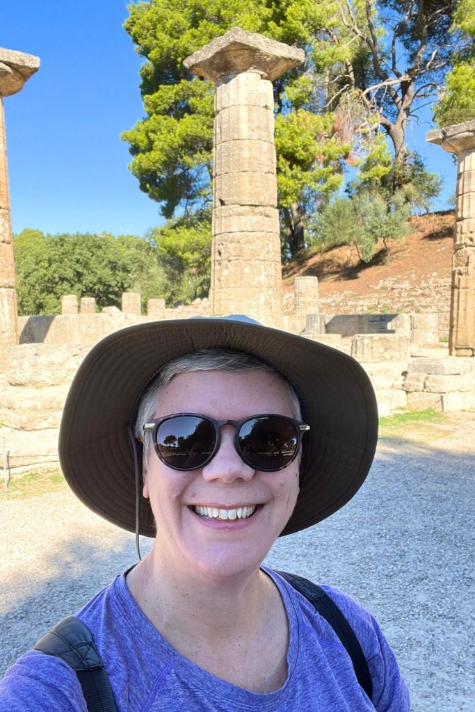 A smiling woman, wearing a purple tee shirt, dark sunglasses, and a wide-brimmed brown hat, stands in front of stone ruins in Ancient Olympia, Greece