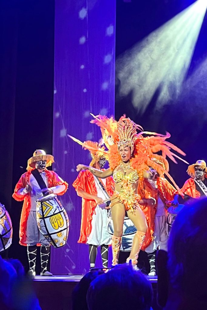 A woman dressed in a carnival costume, with an elaborate headdress and a cape of orange feathers, dances on a stage with four men behind her. The men are also dressed in costumes of white, orange, and yellow, and each is playing a drum that is slung over their chest with a black strap.