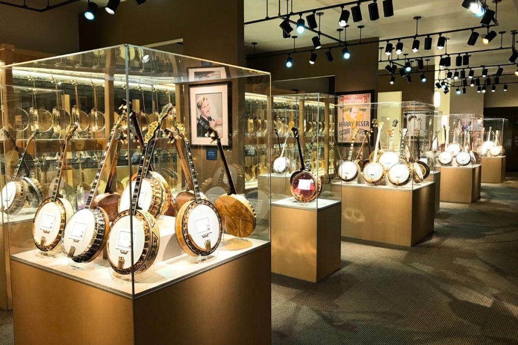 A series of five glass-walled display cases, set in a staggered row, with each display case containing several different banjos set on display stands