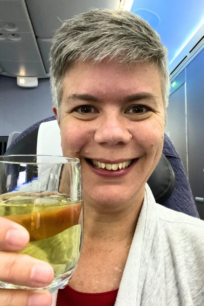 A smiling woman, wearing a gray sweatshirt, holds up a glass of champagne in a toast to the camera, while sitting in an airplane seat
