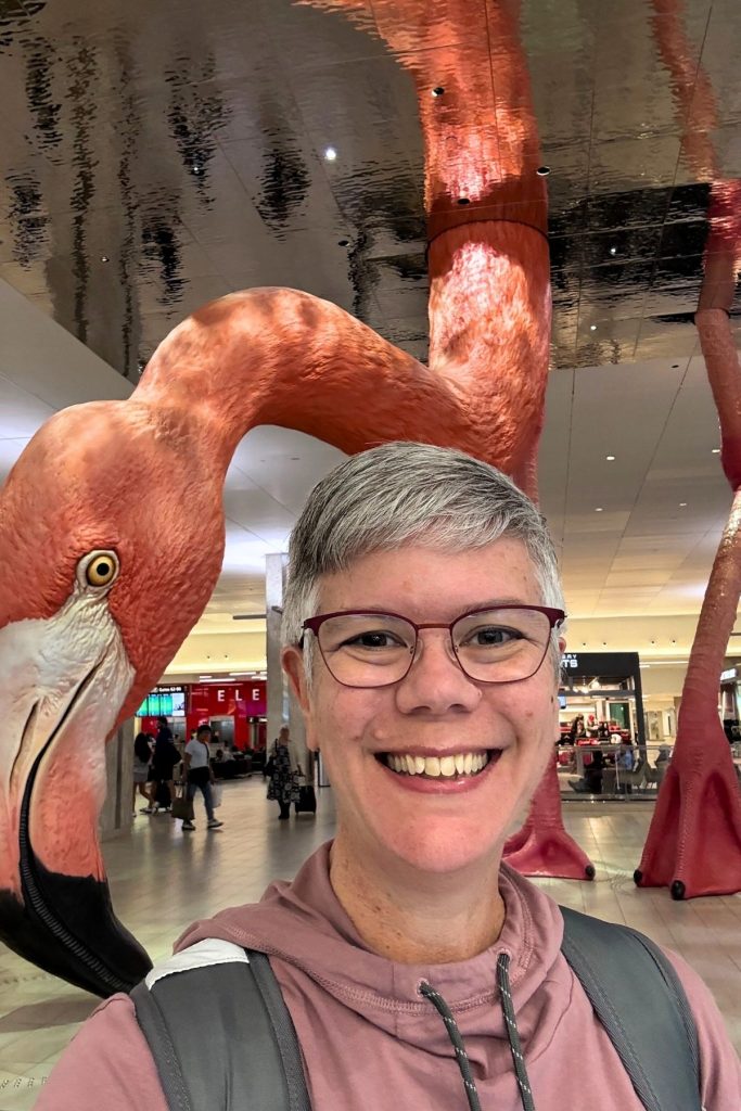 A smiling woman, wearing a mauve colored hooded sweatshirt and eyeglasses, is standing in front of a large statue of a pink flamingo in an airport