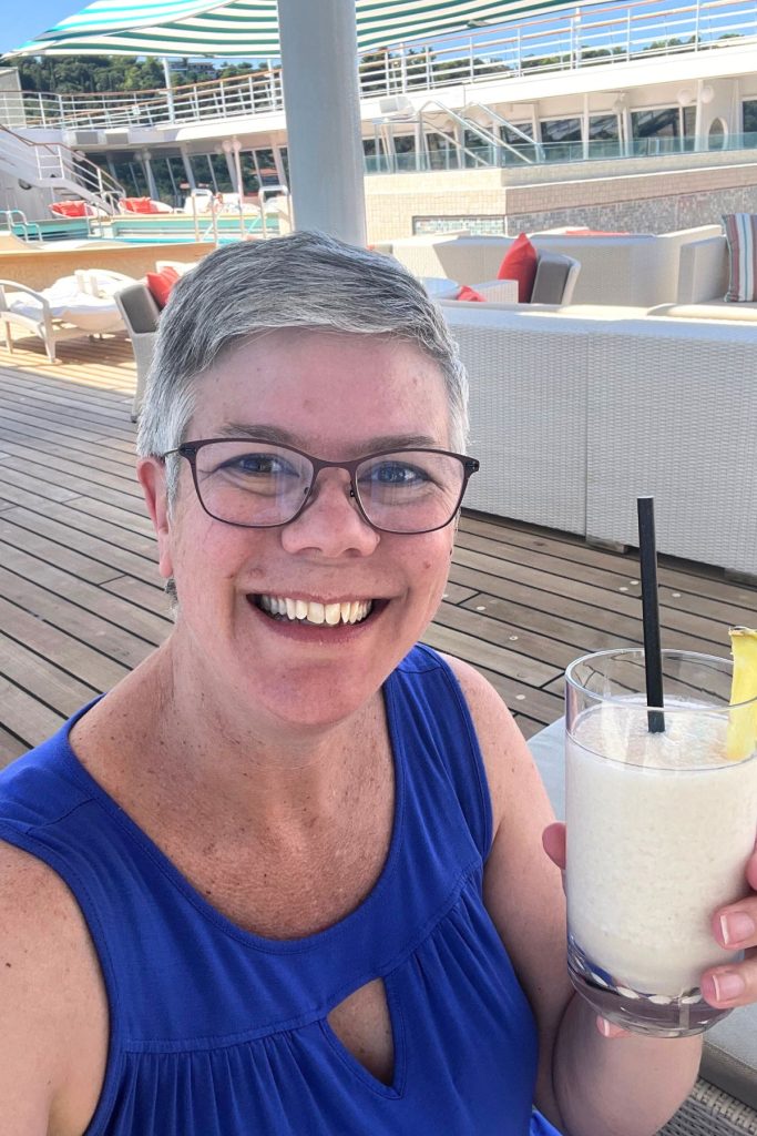 A smiling woman, wearing eyeglasses and a blue tank top, holds a glass of a white frosty drink with a black straw up to the camera in a toasting gesture. She is sitting on the pool deck of a cruise ship.
