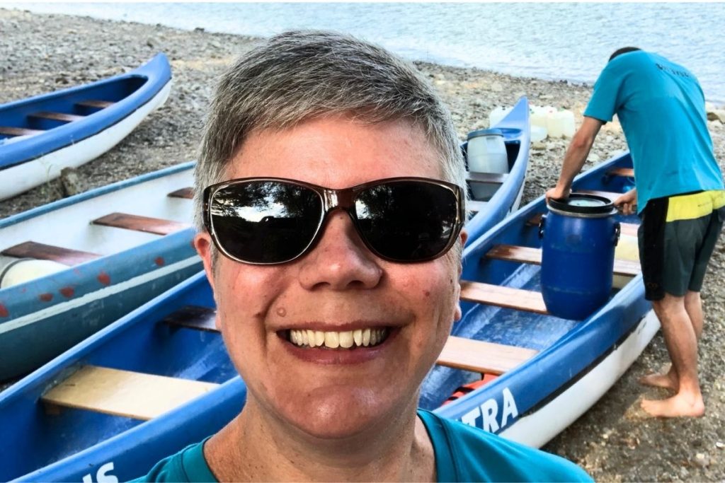 A smiling woman, wearing a teal colored tee shirt and dark sunglasses, stands in front of four blue and white canoes sitting on the shore of a river. A man stands behind her, putting a blue bucket into one of the canoes.
