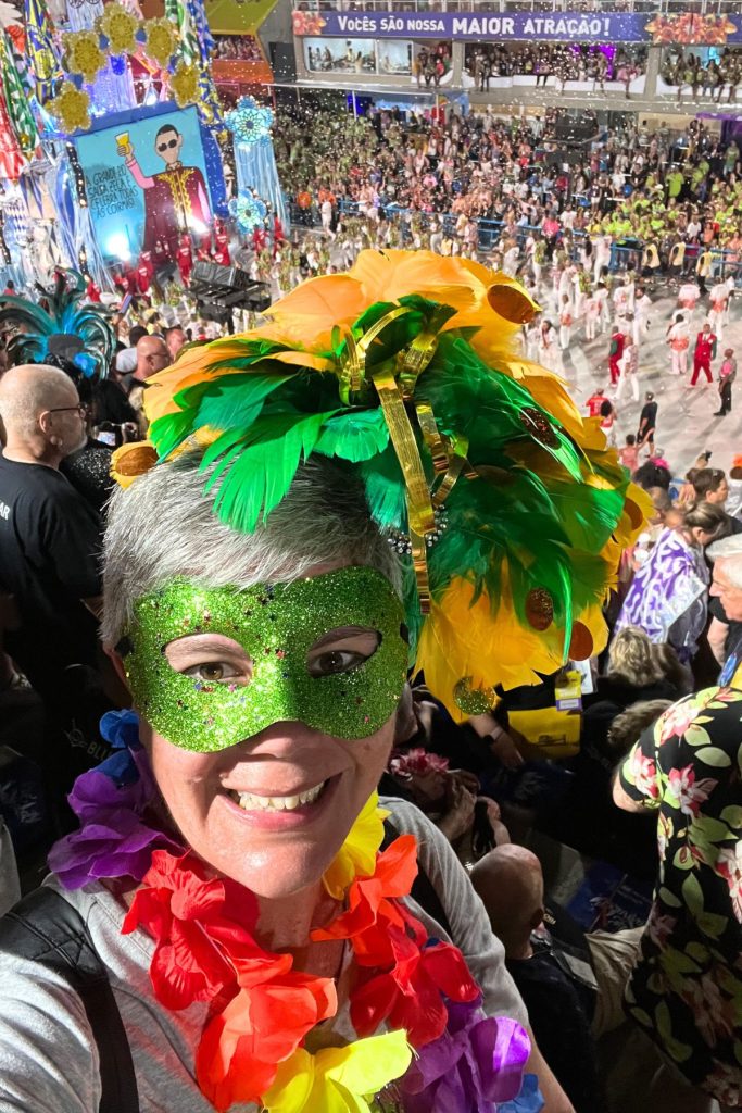 A smiling woman stands in a very large crowd, overlooking a brightly colored Carnival Samba Parade float and performers. The woman is wearing an elaborate headdress with green and yellow feathers, a sparkly green eye mask, and a colorful flower lei around her neck.