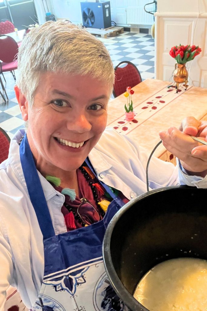 A smiling woman wearing a white button down shirt and a blue apron, holds up a black bucket filled about one third with a white liquid. There is a black and white checkered linoleum floor in the background, with a table covered in a beige cloth decorated with small red flowers, red upholstered chairs, and a bouquet of flowers on the table.