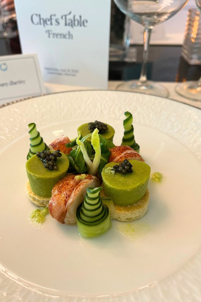 A white plate is artfully arranged with three pieces of cooked lobster meat and three cylinders of green avocado mousse topped with caviar and sitting atop round beige crackers. Three spirals of thinly sliced cucumbers are placed around the plate, along with some leafy greens in the center of the plate.