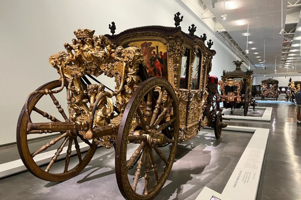Three very ornately decorated and antique horse-drawn carriages sit in a row on white platforms in a museum. Each carriage is decorated with gold carvings and paintings.