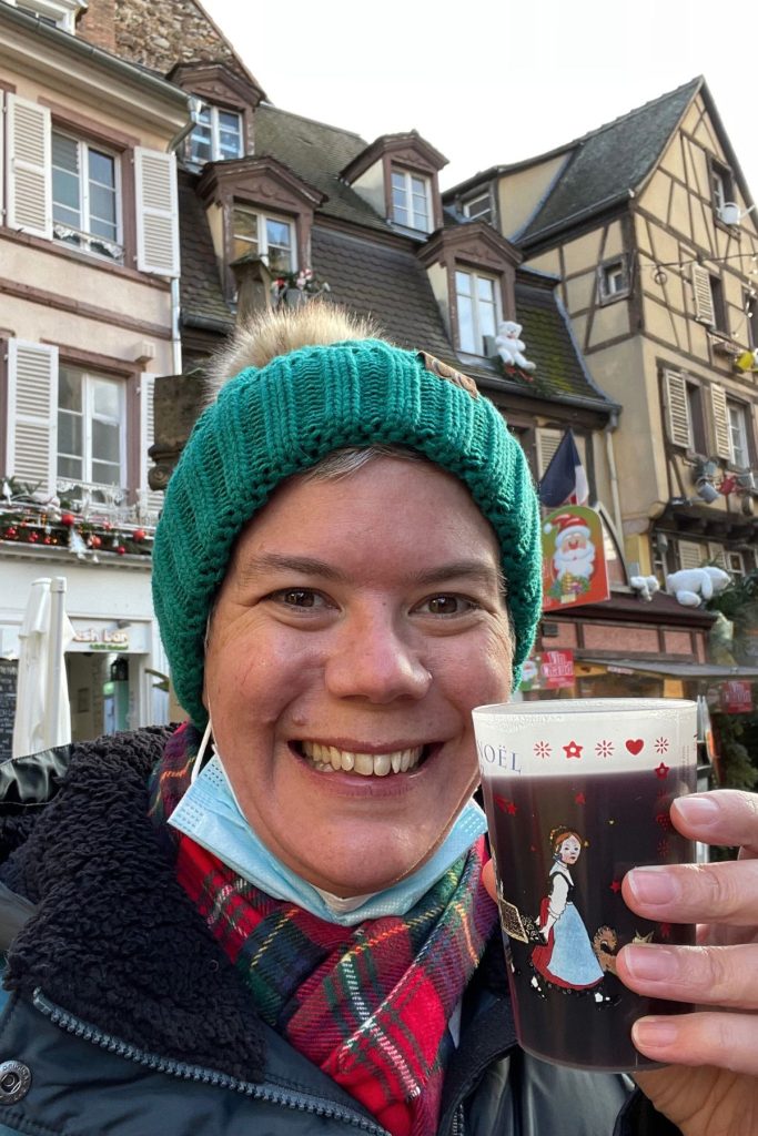 A smiling woman, wearing a green beanie hat, black jacket, and red plaid scarf, holds a cup of mulled wine decorated with cartoon drawings of a French woman at a Christmas market in Colmar, France