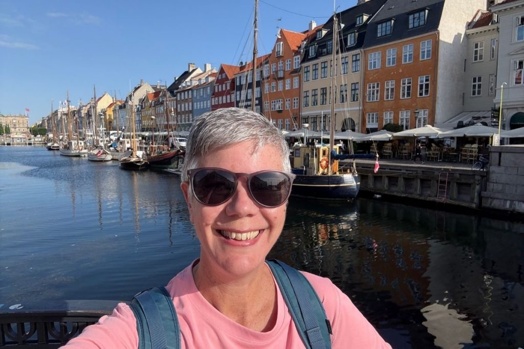 A smiling woman, wearing a pink shirt and dark sunglasses, is standing in front of a row of colorful buildings at Nyhavn in Copenhagen, Denmark