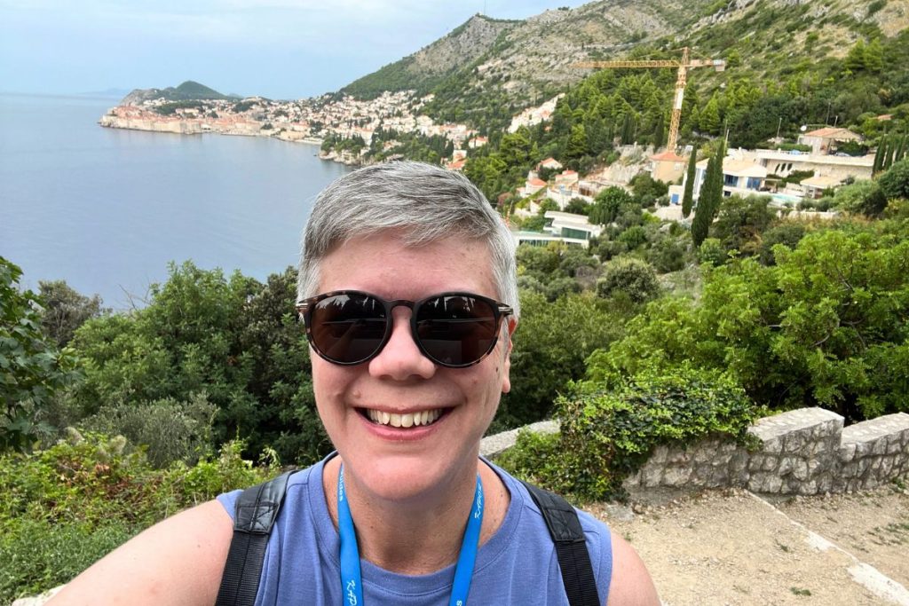 A smiling woman, wearing a purple tank top, dark sunglasses, and a blue lanyard around her neck, is standing on an elevated overlook area, with the city and harbor of Dubrovnik, Croatia behind her. The foreground has many lush green trees and bushes. 