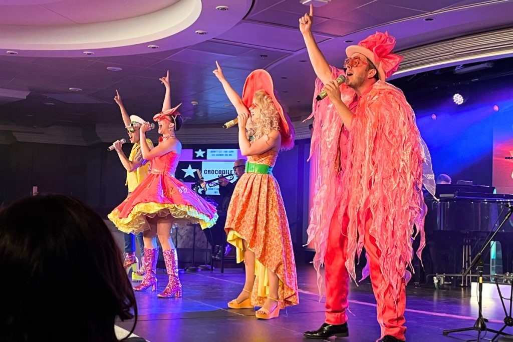 A group of four performers, two men and two women, stand on a stage. Each is dressed in an elaborate costume in shades of oranges, yellows, and greens. Each is singing into a microphone in their left hand, with their right arm extended above their head and their right index finger extended up to the ceiling.