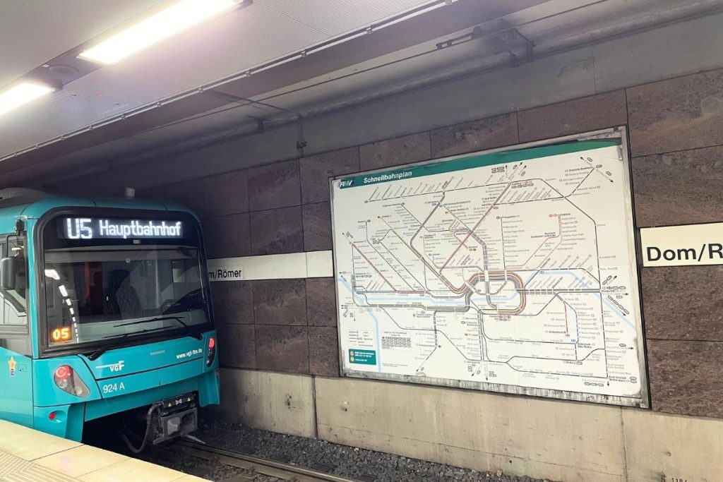 An underground subway stop in Frankfurt, Germany. A teal colored subway train arrives on a track, with a large map of the subway system on the brick wall behind it.