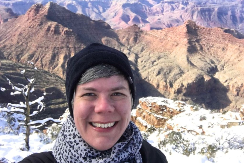 A smiling woman, wearing a black jacket, a black beanie hat, and a black and gray leopard print scarf, stands in front of the Grand Canyon, with some snow seen on the ground directly behind her.