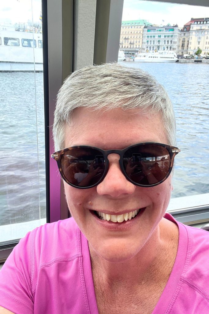 A smiling woman, wearing a bright pink shirt and dark sunglasses, sits inside an enclosed boat on a river, with the river seen directly behind the windows behind the woman, A few buildings and boats are seen in the background.