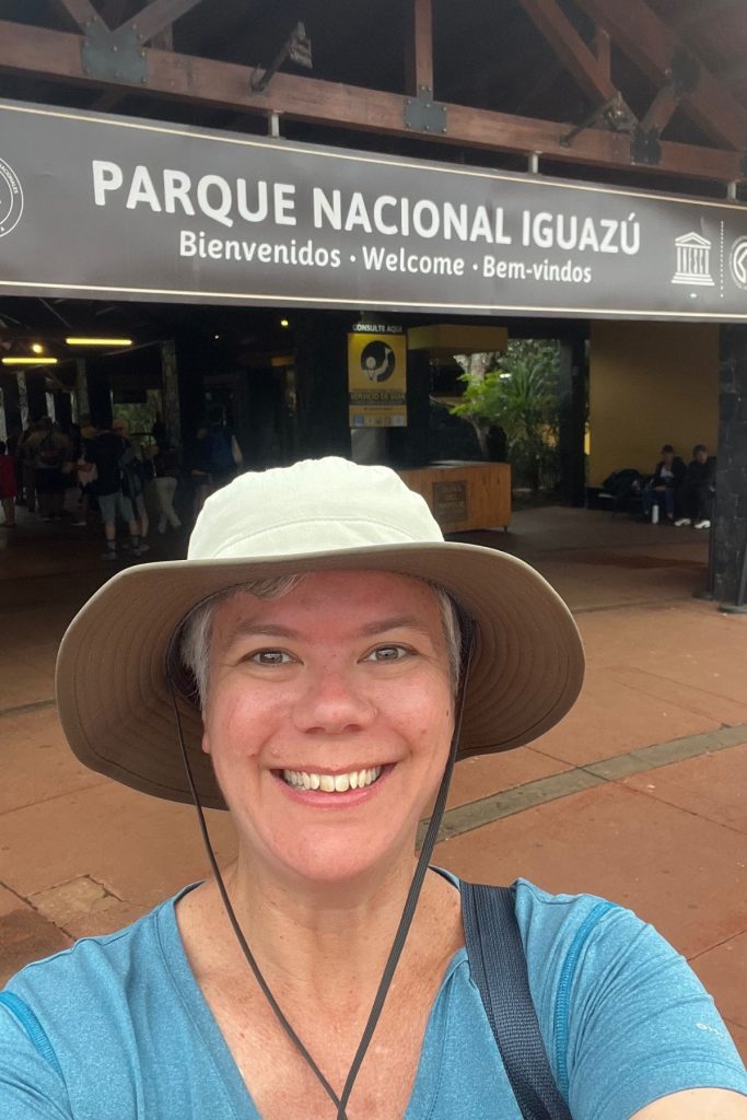 A woman in a tan bucket hat smiling in front of the Parque Nacional Iguazú entrance sign