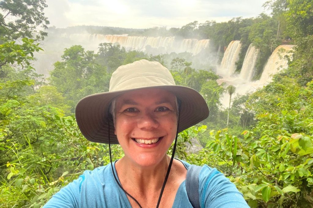 A woman in a tan bucket hat smiling in the rainforest with a line of falls in the mist behind her