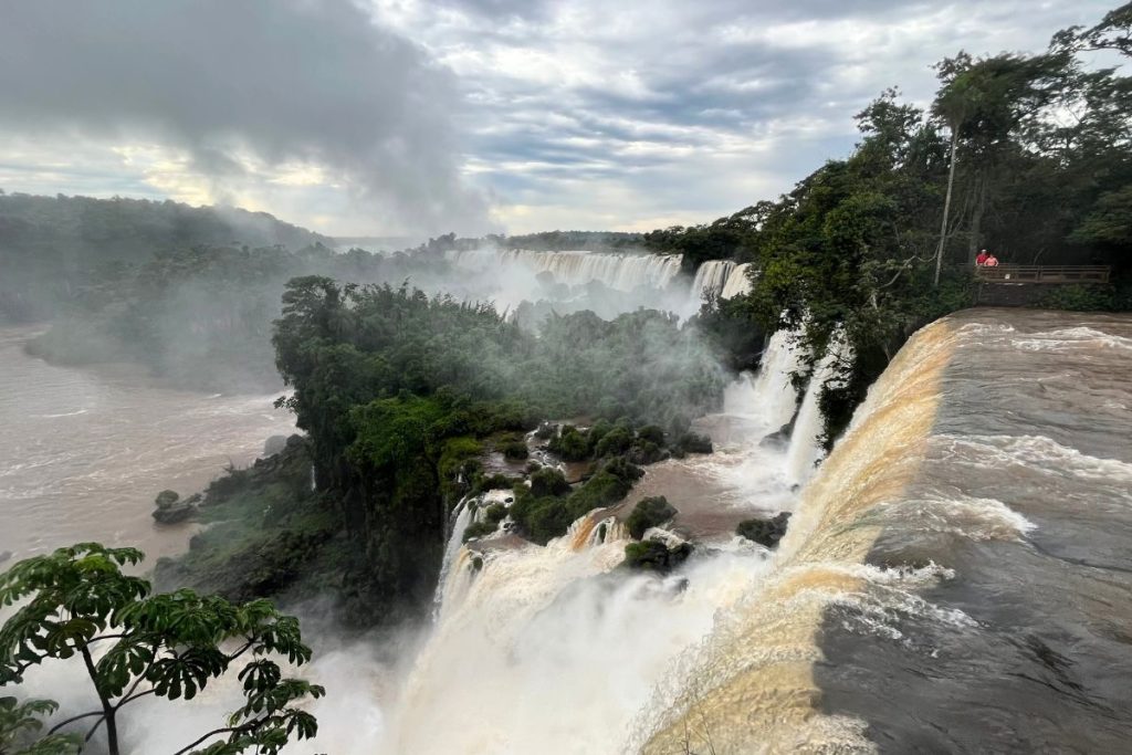 River rushing over the curved edge of the falls, with forested islands and misty falls beyond