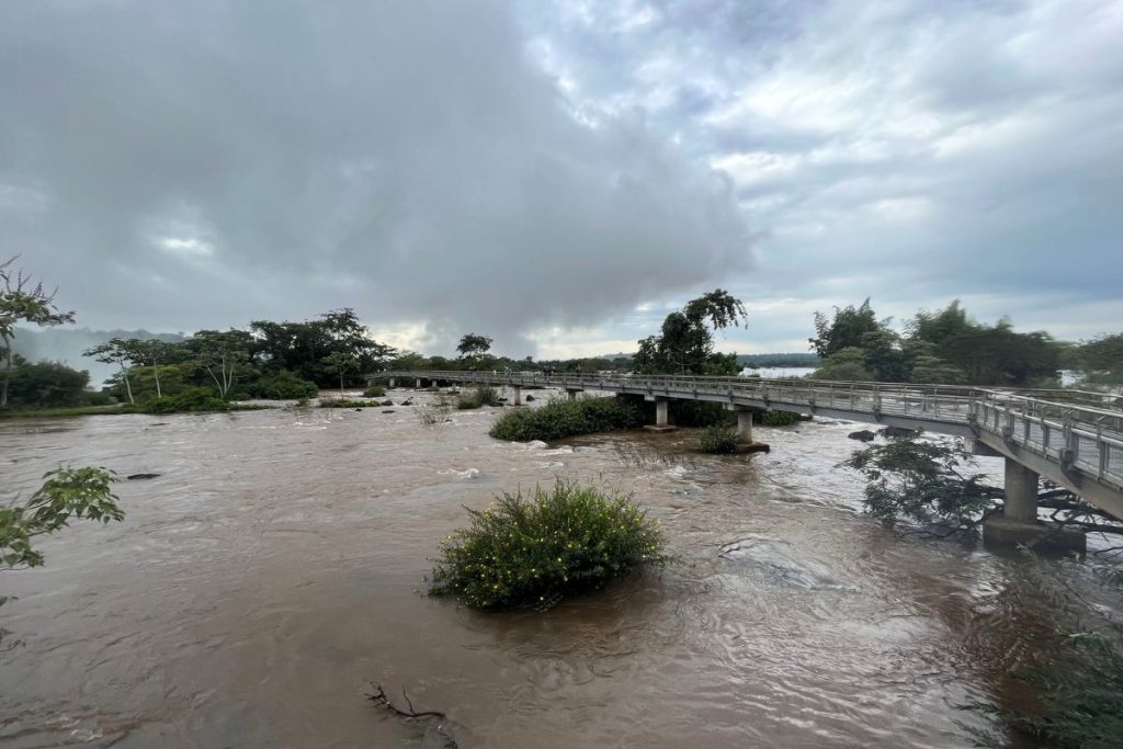 Long concrete walkway crossing a wide, fast brown river under heavy gray clouds