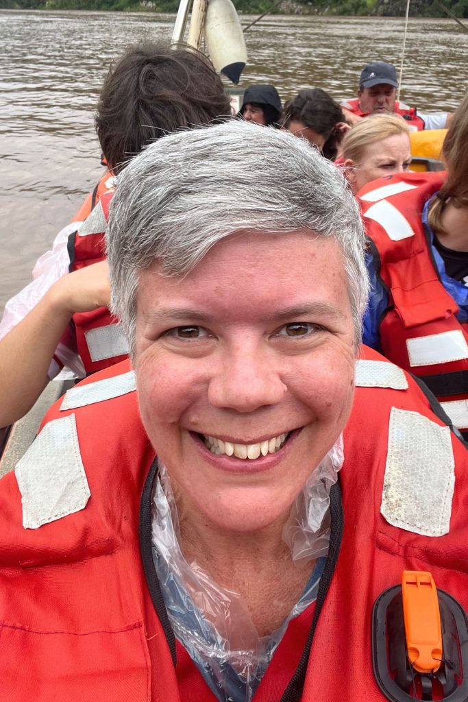 A woman smiling in a red life jacket with dry gray hair aboard a boat, other passengers behind her