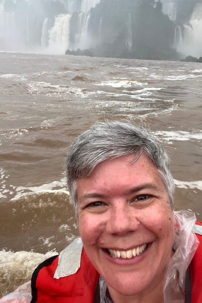 A woman in a red life jacket and plastic poncho on a boat, with falls in the mist behind her