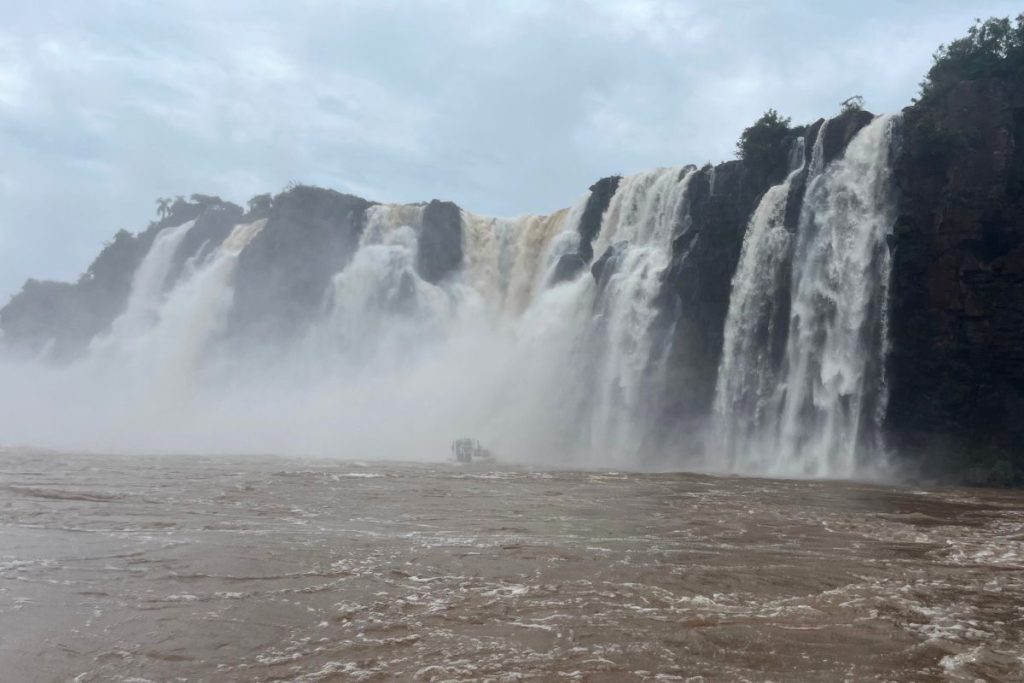 Tall wall of falls spilling over dark cliffs into a brown river, with a small boat in the mist