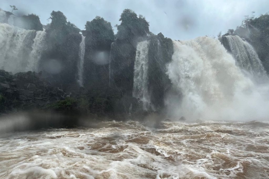 Falls crashing over dark rock into churning brown water, with raindrops on the camera lens