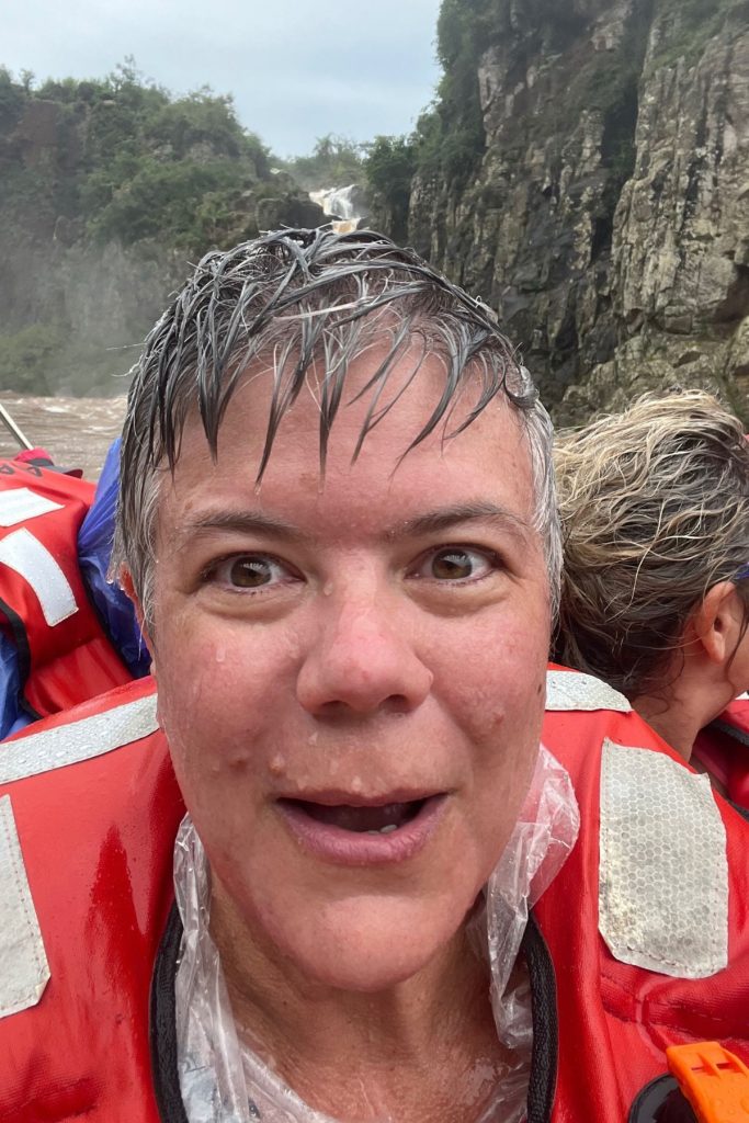 A woman with soaked, spiked hair and wide eyes in a red life jacket, a rock cliff behind her