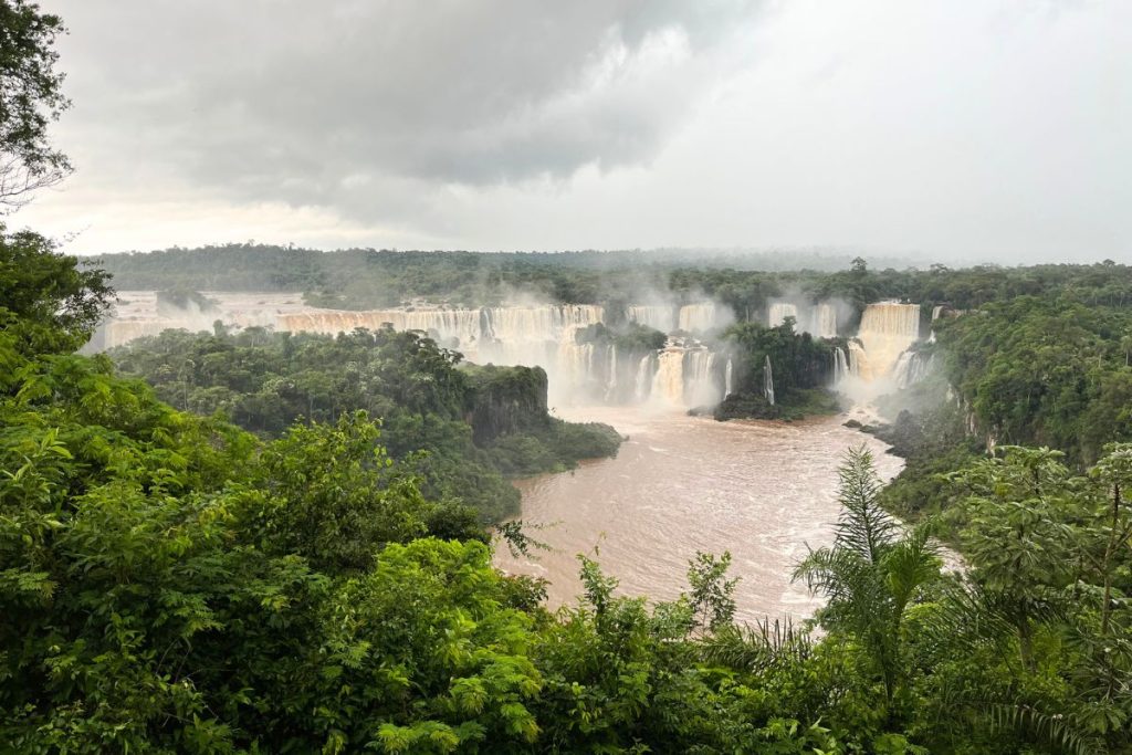Iguazu Falls seen through rainforest foliage under gray clouds, with a brown river below