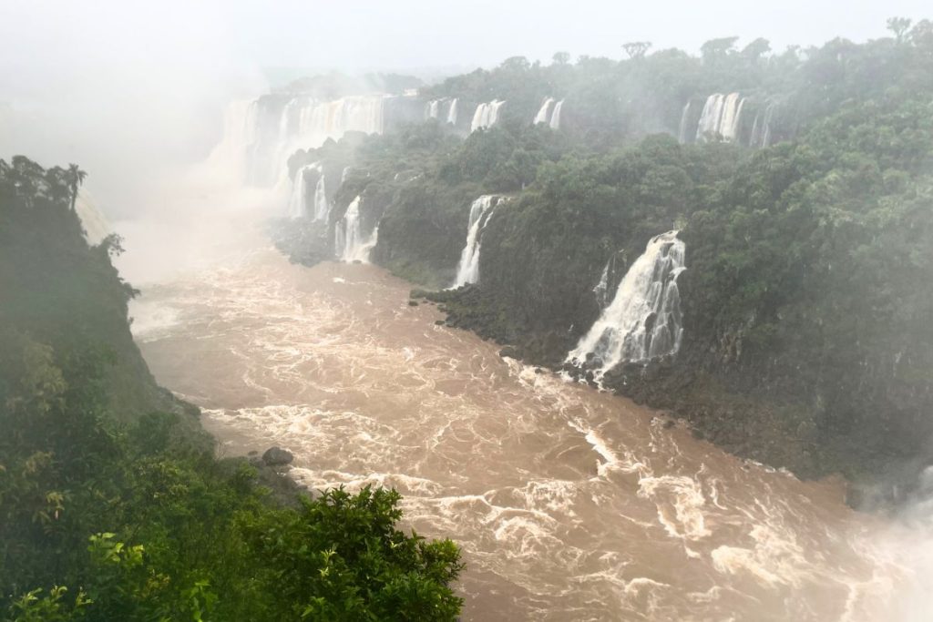Brown rapids in a forested canyon with several waterfalls dropping from the cliffs and mist rising