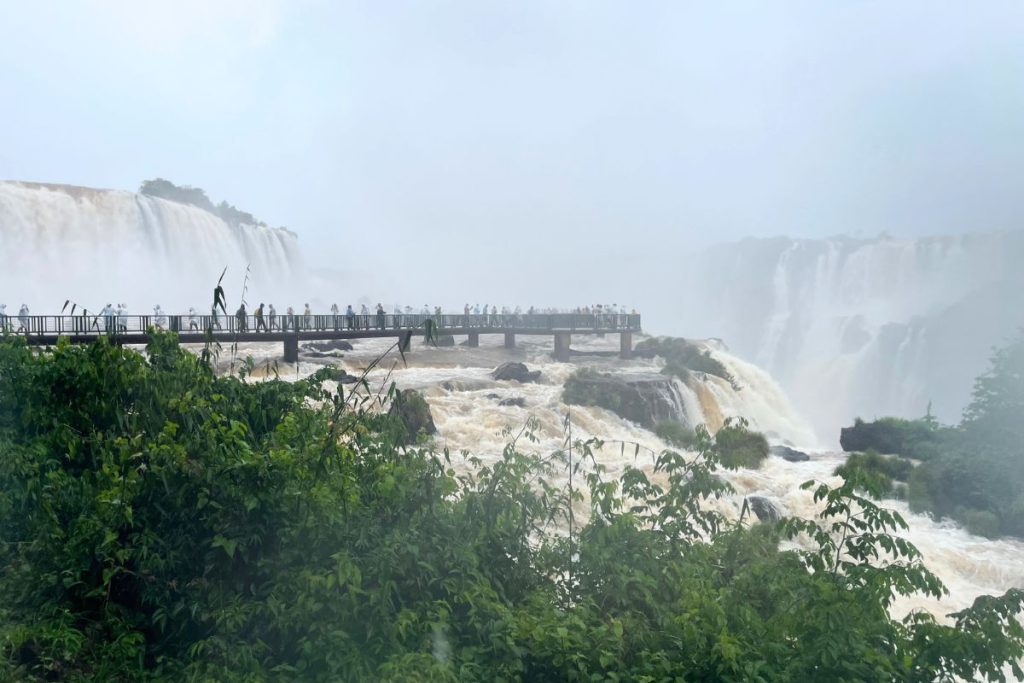 Visitors in ponchos on a walkway platform over brown rapids, with a large waterfall in the mist behind