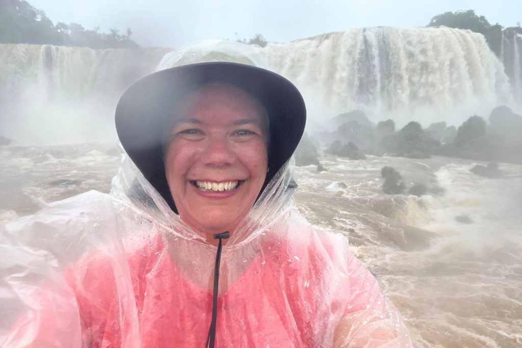 A woman in a bucket hat and clear poncho, smiling, with misty falls and brown water behind her