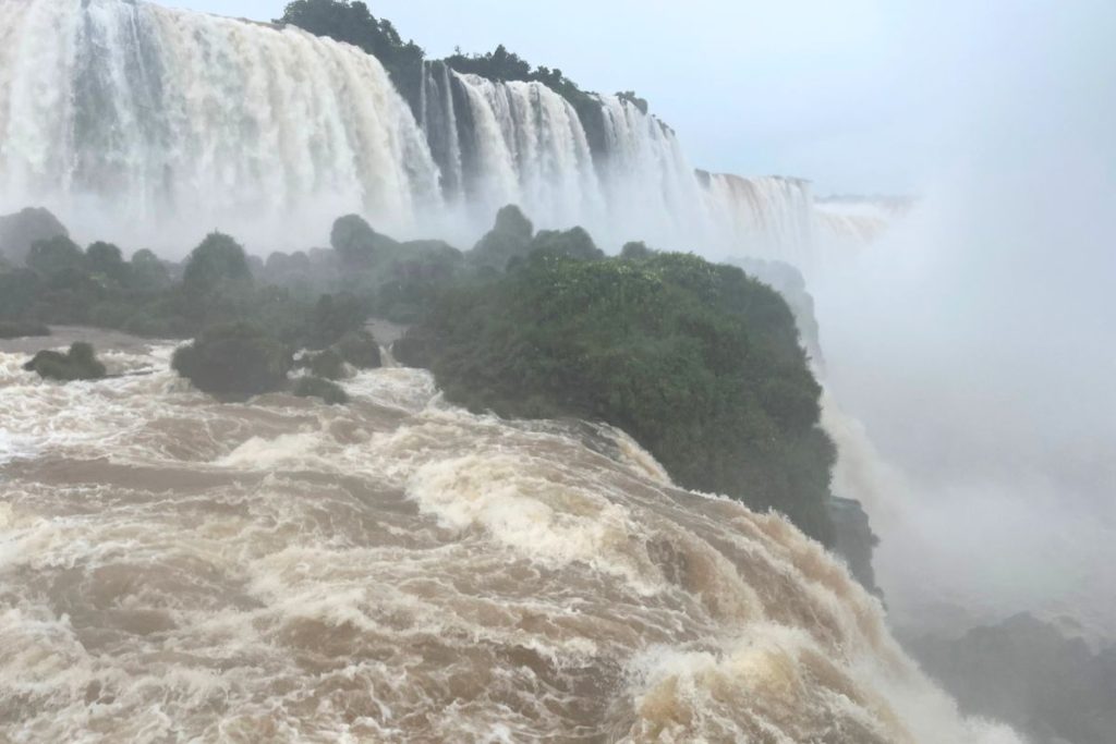Brown rapids rushing toward the edge of the falls, with a tall misty waterfall beyond a green island