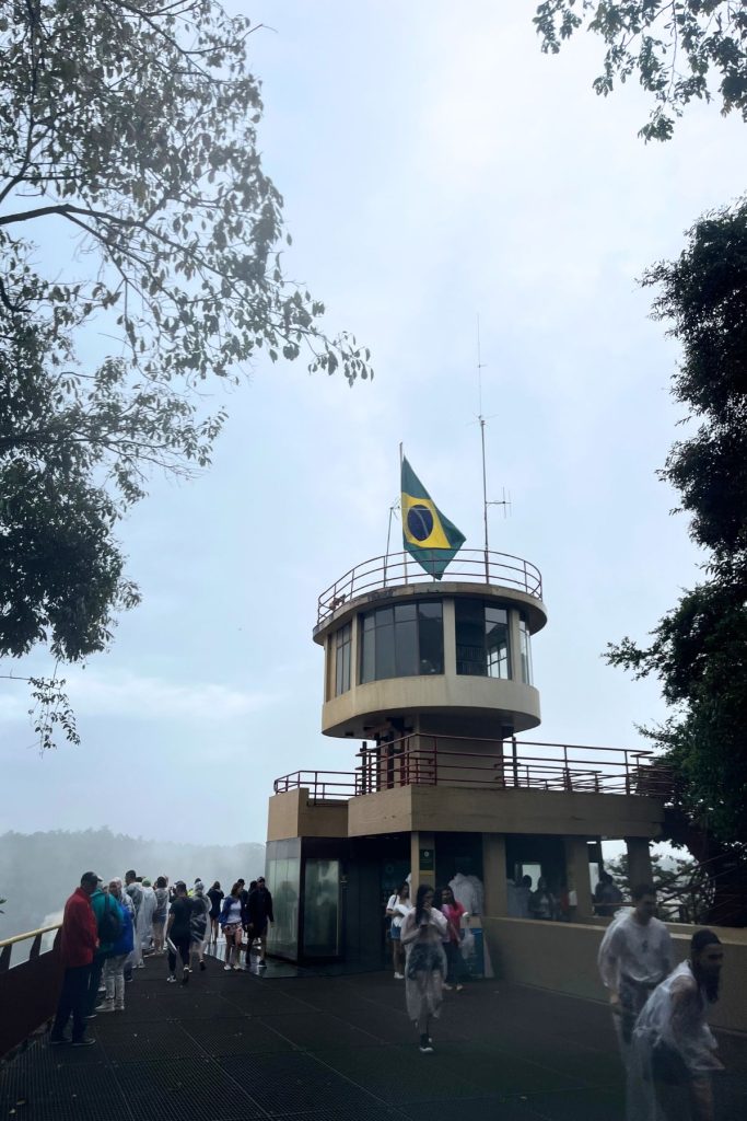 Round tower with a Brazilian flag above a wet viewing platform with visitors in clear ponchos