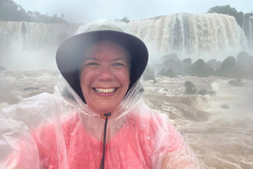A smiling woman, wearing a red shirt with a clear rain poncho on top and a wide brimmed hat, stands in front of a very large waterfall