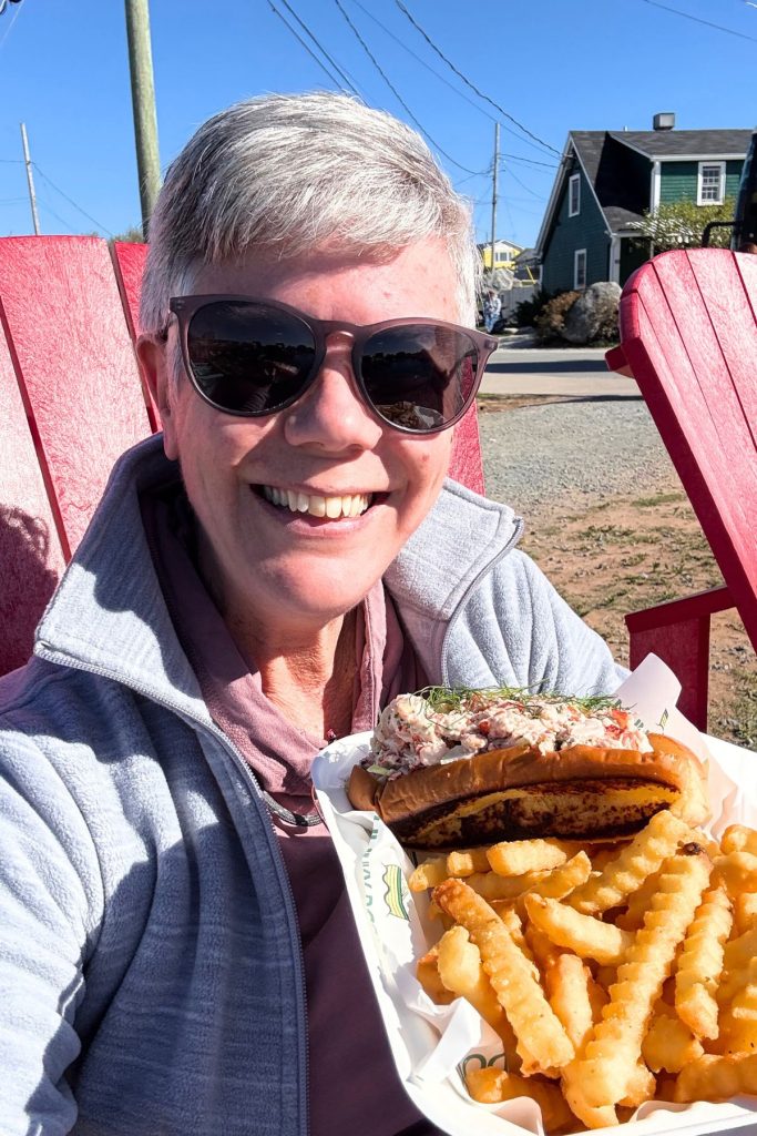 A smiling woman, wearing a gray fleece jacket, pink hooded sweatshirt, and dark sunglasses, sits on a red Adirondack chair with a green house in the background. The woman holds up a plate with a lobster roll and French fries.