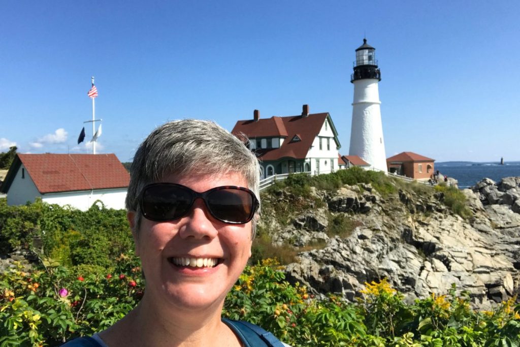 A smiling woman wearing dark sunglasses stands in front of the Portland Head Light, a famous lighthouse in Portland, Maine.