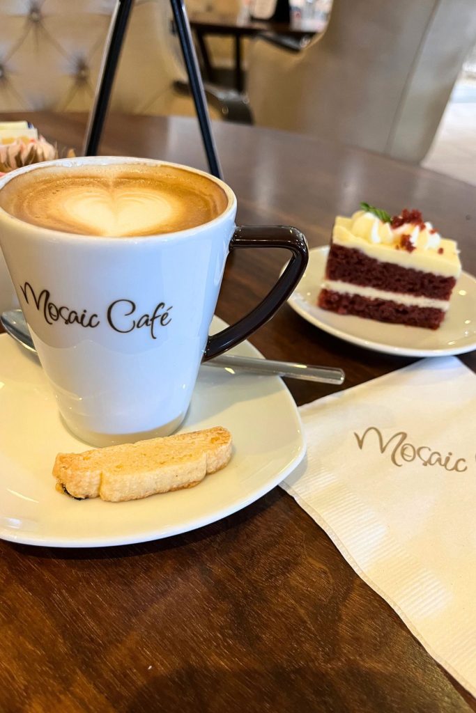 A cup of cappuccino in a white coffee mug on a white saucer, with the words Mosaic Cafe in black on the front of the mug. The saucer has a spoon resting on it behind the mug, and a small piece of biscotti cookie resting on it in front of the mug. A piece of red velvet cake on a white plate is behind the mug. 