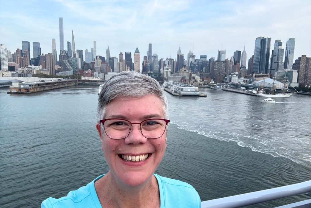 A smiling woman, wearing a turquoise colored shirt and eye glasses, stands on the deck of a cruise ship, with the skyline of New York City in the background