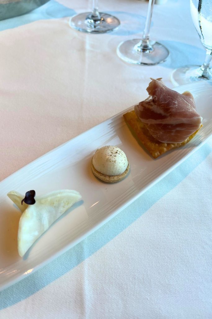 An oblong white plate with three small appetizers placed on top sits on a table covered in a white cloth. Stems of wine glasses are seen in the background.