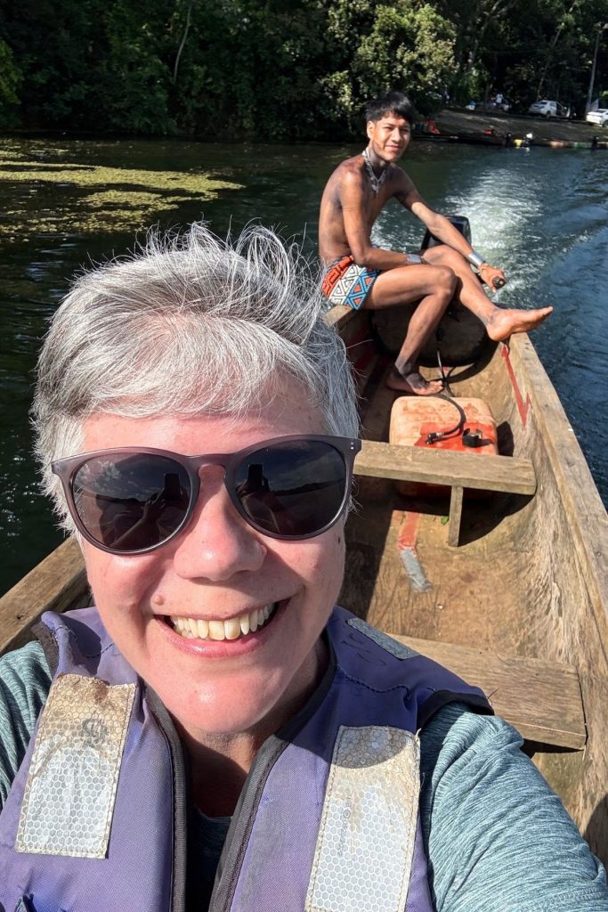 A smiling woman, wearing a purple life jacket and dark sunglasses, sits in a primitive wooden canoe, with a native villager sitting at the back and steering the canoe