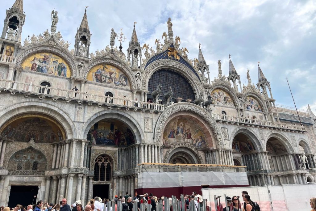 The front entrance of Basilica San Marco in Venice, Italy. The top is adorned with several carved figures, arranged on the top of a series of arches with paintings set in them. The bottom row of arches cover windows and open doorways, with a long line of people waiting to enter seen at the bottom of the picture.