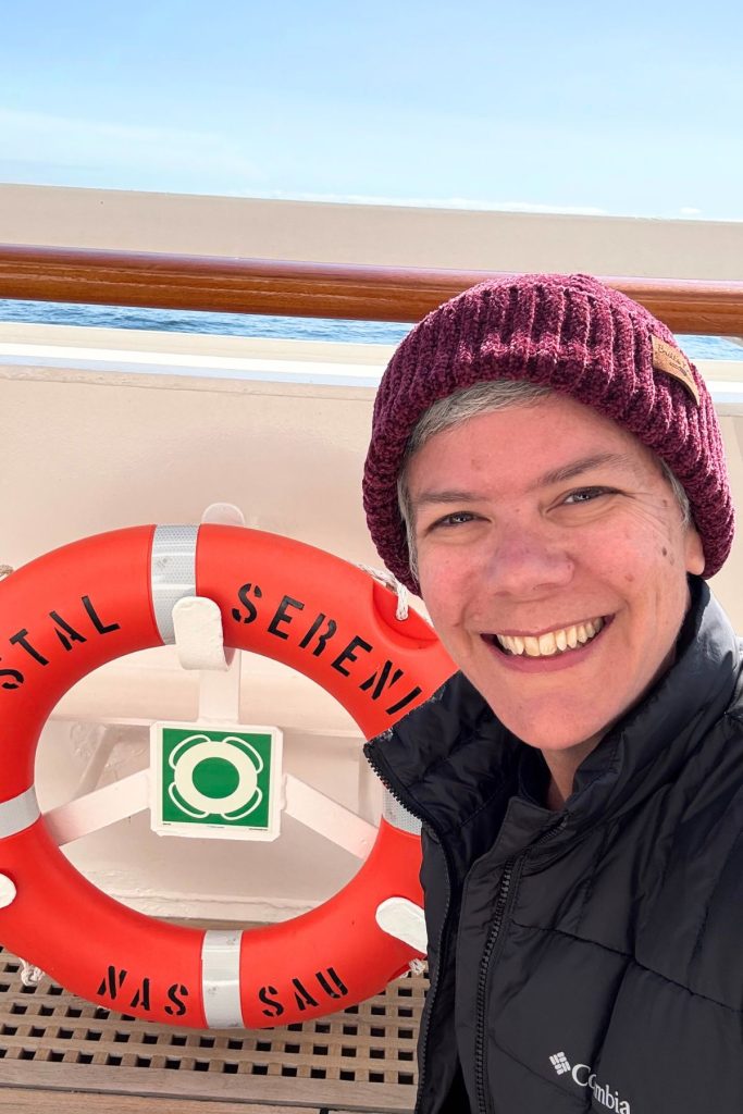 A smiling woman, wearing a black coat and a maroon beanie hat, crouches next to an orange life saving ring on a cruise ship deck. The ring has the words Crystal Serenity in black on the top, and the word Nassau in black on the bottom. A small sliver of water can be seen behind the woman's head.
