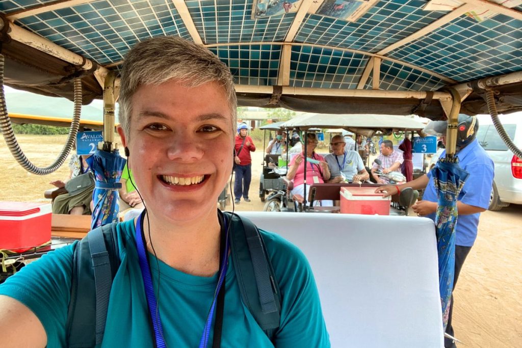 Smiling woman in a teal shirt taking a selfie in a tuk-tuk, with other Avalon Waterways tuk-tuks and passengers behind her.
