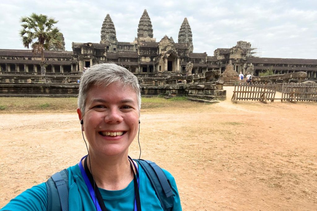 Woman in a teal shirt smiling in front of Angkor Wat’s towers and outer gallery, with a palm tree at left.