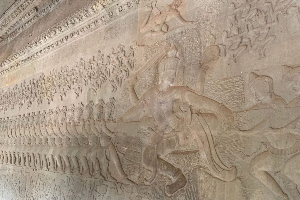 Stone bas-relief with a multi-armed Vishnu at center and a line of figures pulling a serpent, with rows of small celestial figures above.