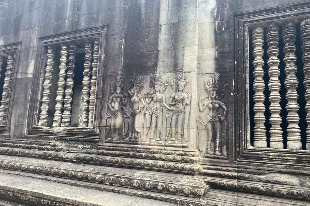 Five carved apsaras with tall crowns standing side by side on a weathered stone wall between two windows with carved balusters.