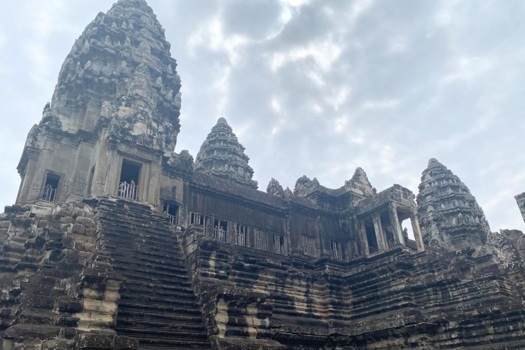 Looking up a steep stone staircase to an Angkor Wat tower under a cloudy sky, with visitors in the upper windows.