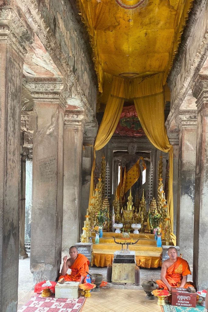 Two monks in orange robes seated beside donation boxes in front of a gold-draped Buddha shrine between stone pillars.