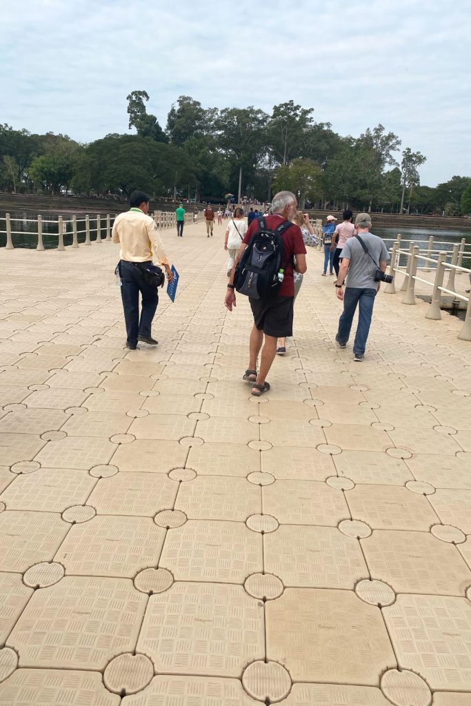 Visitors walking across a floating bridge of linked beige tiles over the moat, with trees ahead.