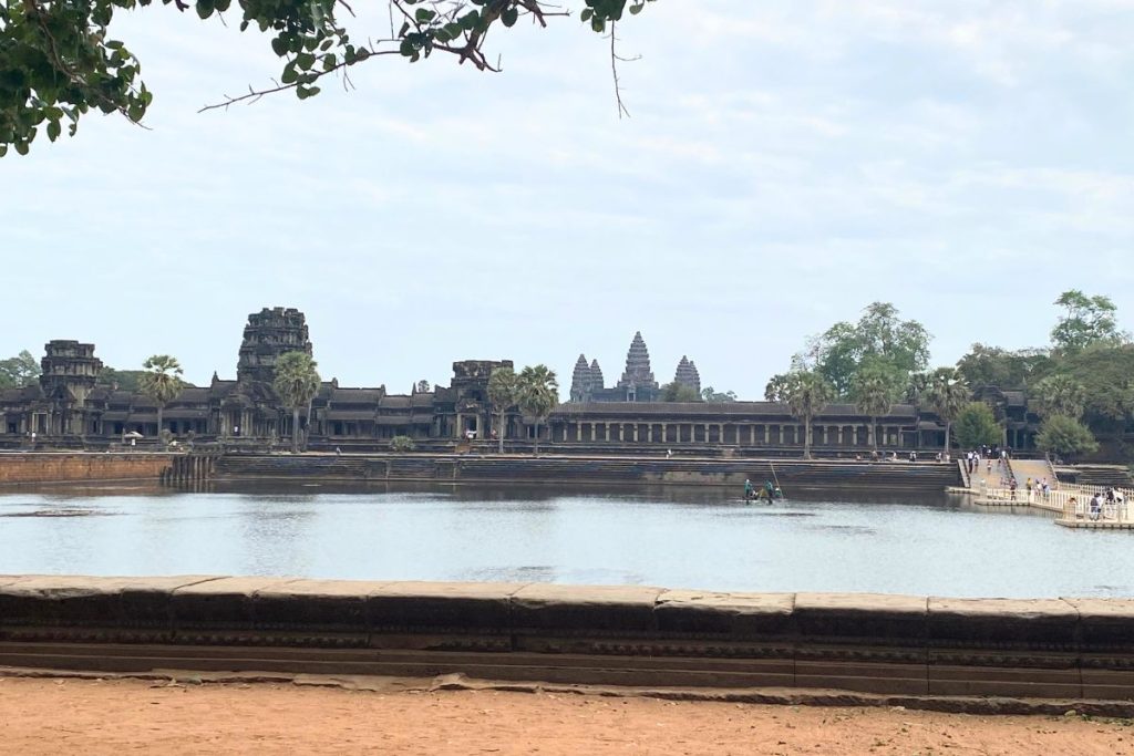 View across the moat to Angkor Wat’s long galleries and three visible towers, with palm trees and a floating bridge at right.